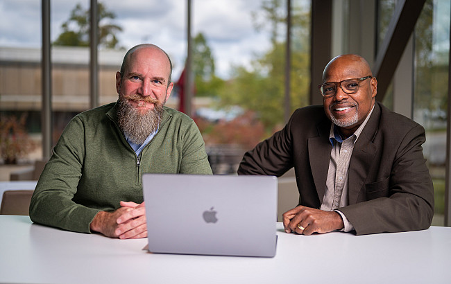 Two men sit a table with a laptop.