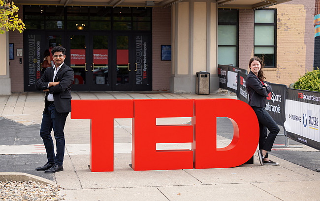 Two Indiana University students lean against the outside edges of a set of large red letters that spell TED in front of a theater entrance