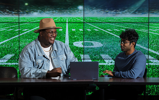 Two students sit talking in front of a laptop with a projection of a football field on the wall behind them.