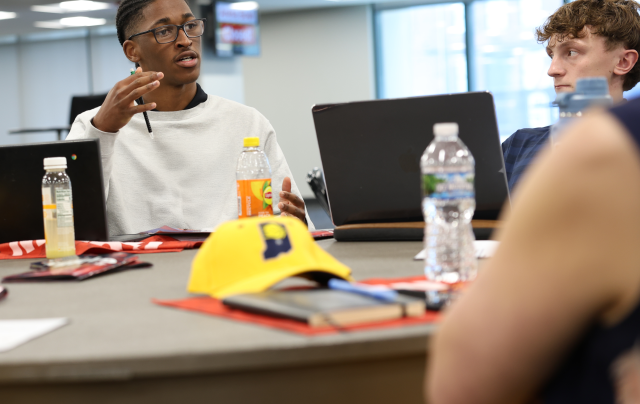 Two students sit at a table and converse. There is a yellow Indiana Pacers hat on the table.
