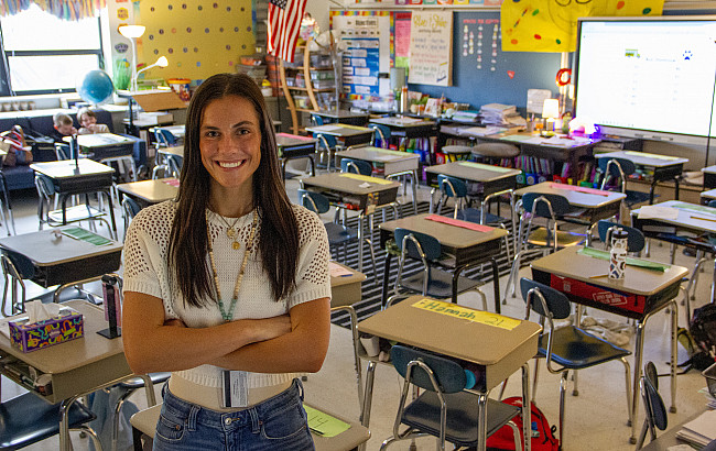 A women stands smiling in front of a classroom full of desks.