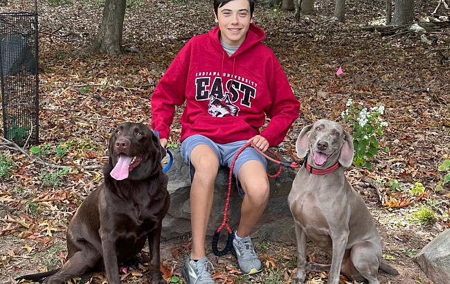 Nathan in an IU East hoodie sitting on a rock in a wooded area with two dogs on leashes at his side