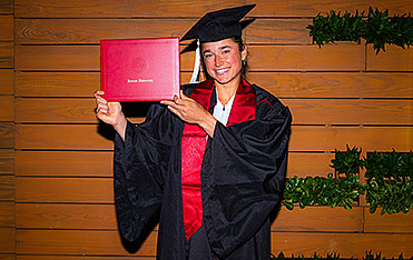 Dolehide smiling in her cap, gown and stole, holding up her red IU degree case