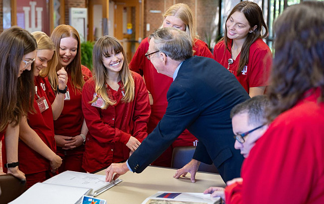 A group of young woment listening to a man in a dark jacket as he points to a scrapbook