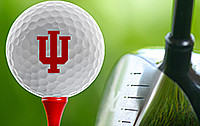 Golf ball with Indiana University trident sitting on a golf tee with golf club next to the ball and tee.