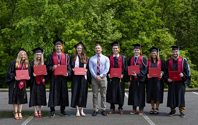 Seven graduates posed together, one in business clothes, the rest in commencement gowns and hats.