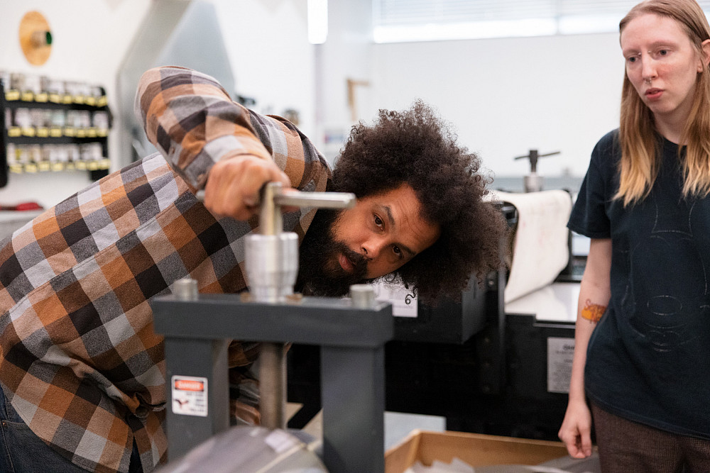 Professor Aaron Coleman leans down behind a piece of printmaking equipment while turning a handle on the top of the machine. 